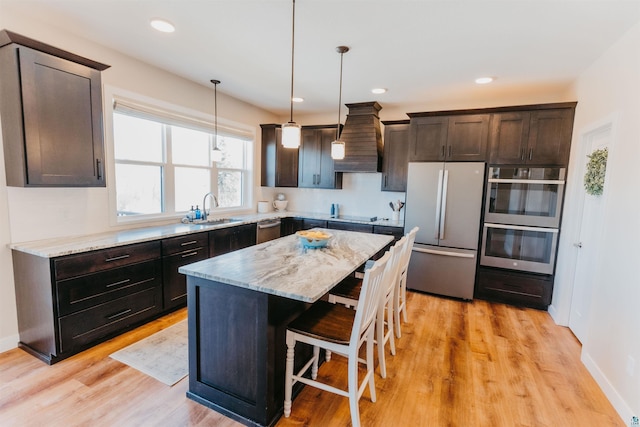 kitchen featuring a sink, stainless steel appliances, custom range hood, dark brown cabinets, and a kitchen bar