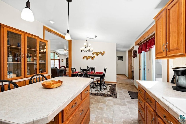 kitchen with ceiling fan with notable chandelier, decorative light fixtures, brown cabinetry, light countertops, and glass insert cabinets