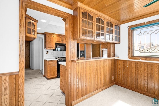 kitchen featuring brown cabinets, black appliances, wooden walls, wainscoting, and glass insert cabinets