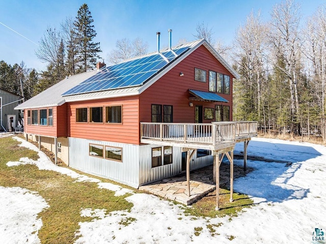 snow covered back of property featuring a deck, a chimney, roof mounted solar panels, and metal roof