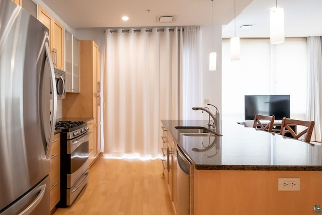 kitchen with an island with sink, dark stone countertops, appliances with stainless steel finishes, light wood-style floors, and a sink