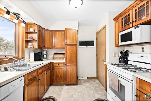 kitchen with a sink, white appliances, brown cabinetry, light countertops, and glass insert cabinets