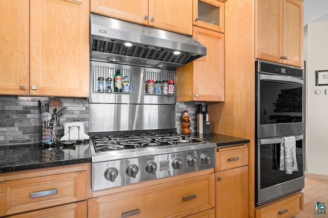 kitchen featuring stainless steel appliances, dark stone counters, backsplash, glass insert cabinets, and under cabinet range hood
