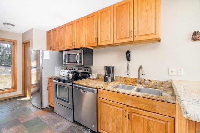 kitchen with stainless steel appliances, a sink, stone finish flooring, light stone countertops, and brown cabinetry