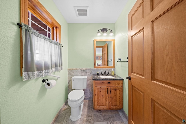 half bath with visible vents, toilet, tile walls, vanity, and wainscoting
