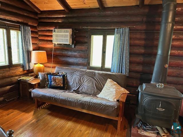 sitting room featuring a wood stove, rustic walls, hardwood / wood-style floors, beamed ceiling, and a wall mounted AC