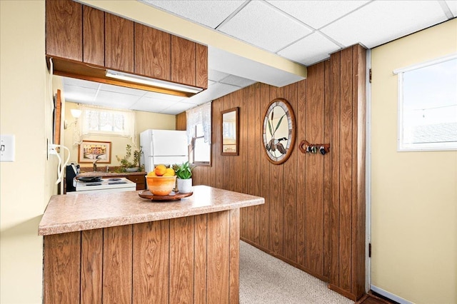 kitchen with wood walls, light carpet, white appliances, a drop ceiling, and brown cabinets