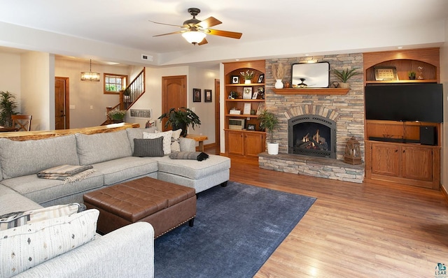 living room with a stone fireplace, visible vents, wood finished floors, ceiling fan with notable chandelier, and stairs