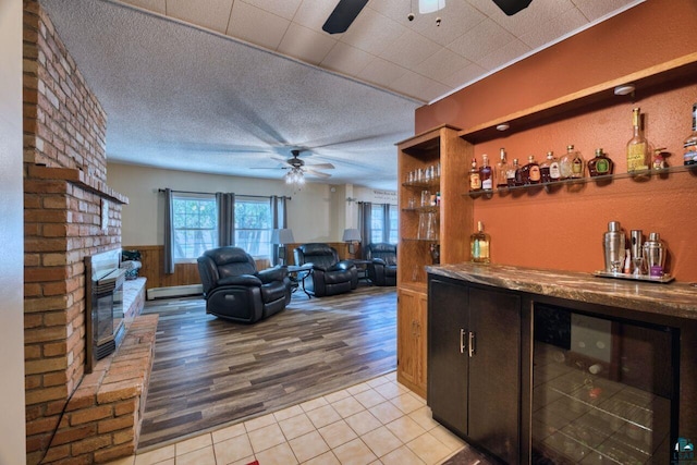 bar featuring a ceiling fan, a bar, a fireplace, a baseboard radiator, and wine cooler