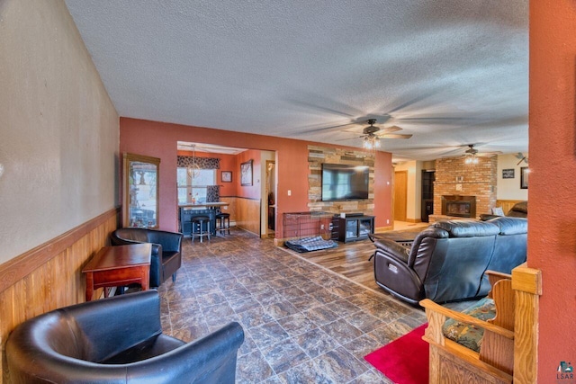 living area with a ceiling fan, a brick fireplace, a textured ceiling, wainscoting, and wooden walls