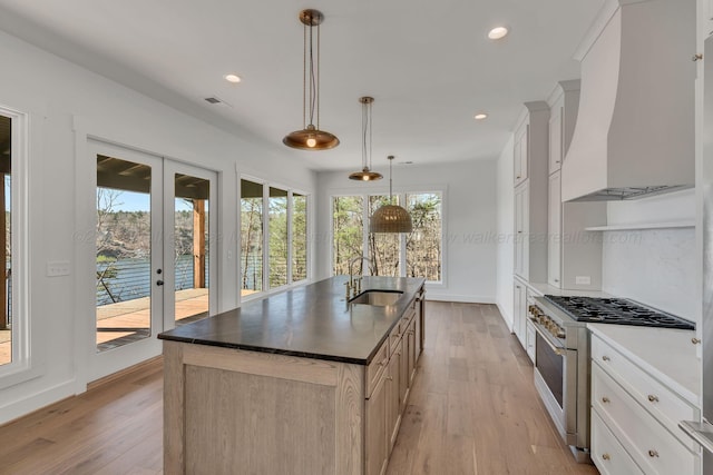 kitchen with a center island with sink, light wood finished floors, custom exhaust hood, dark countertops, and gas range