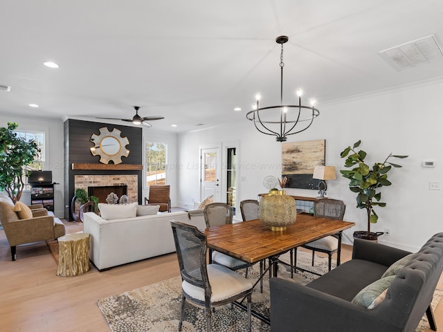 dining room featuring a stone fireplace, crown molding, ceiling fan with notable chandelier, and light wood-type flooring