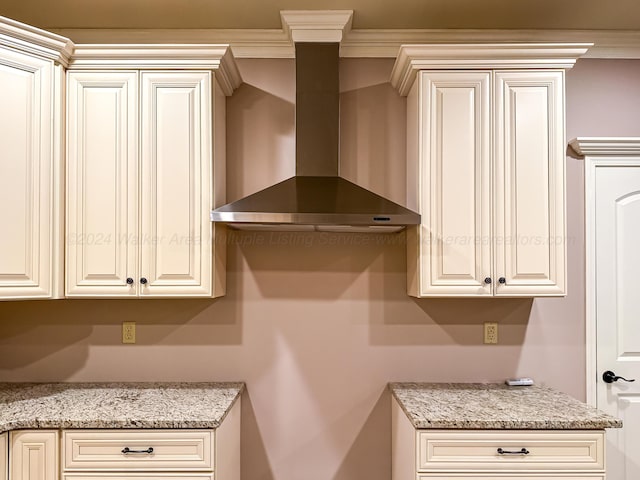 kitchen with light stone countertops, cream cabinetry, ornamental molding, and wall chimney range hood