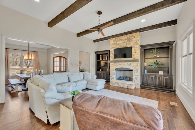 living room with ceiling fan with notable chandelier, a stone fireplace, beamed ceiling, and wood finished floors