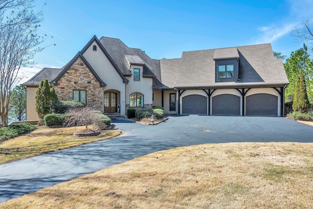 french country inspired facade with an attached garage, driveway, stone siding, stucco siding, and a front yard