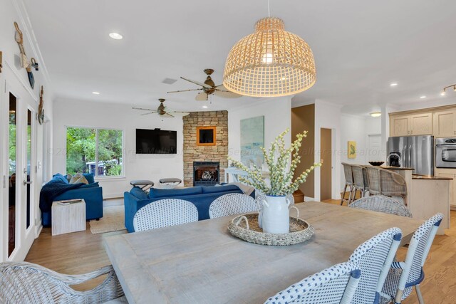 dining room with ceiling fan, light hardwood / wood-style floors, a stone fireplace, and ornamental molding