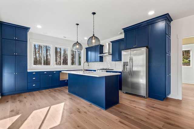 kitchen with blue cabinets, wall chimney range hood, and stainless steel fridge with ice dispenser