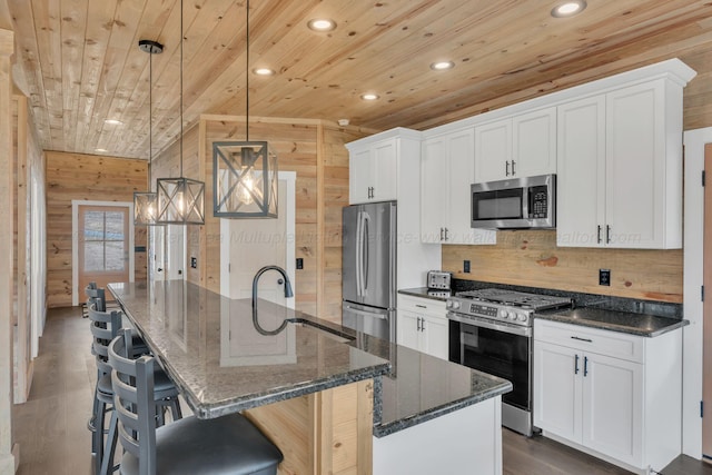 kitchen with wooden walls, an island with sink, wood ceiling, stainless steel appliances, and a sink