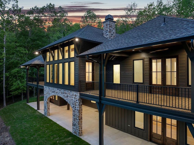 property exterior at dusk with roof with shingles, a patio area, stone siding, a yard, and a chimney