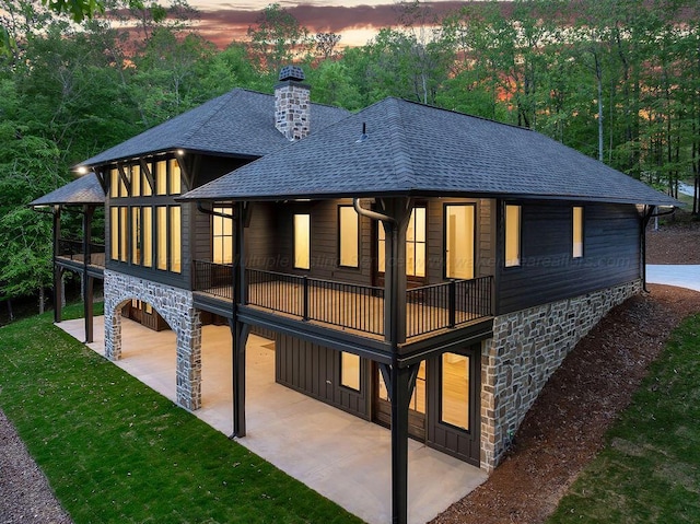 rear view of house featuring a chimney, a yard, a patio area, stone siding, and roof with shingles