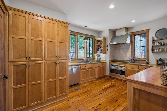 kitchen with appliances with stainless steel finishes, light wood-type flooring, sink, wall chimney range hood, and hanging light fixtures