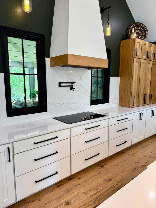 kitchen with black electric cooktop, tasteful backsplash, light wood-style floors, white cabinetry, and custom range hood