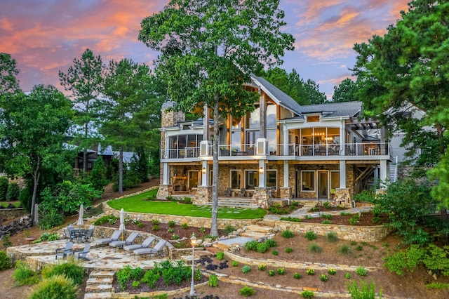 back of property at dusk with stone siding, a sunroom, a wooden deck, stairs, and a patio area