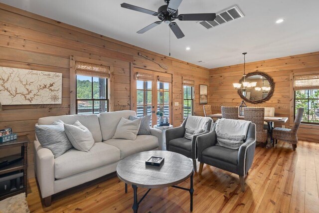 living room featuring ceiling fan with notable chandelier, wooden walls, light hardwood / wood-style flooring, and a healthy amount of sunlight