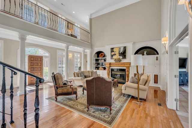 living room with a high ceiling, crown molding, built in shelves, light wood-type flooring, and ornate columns