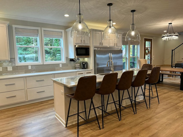 kitchen featuring built in appliances, white cabinetry, light wood-type flooring, pendant lighting, and an island with sink