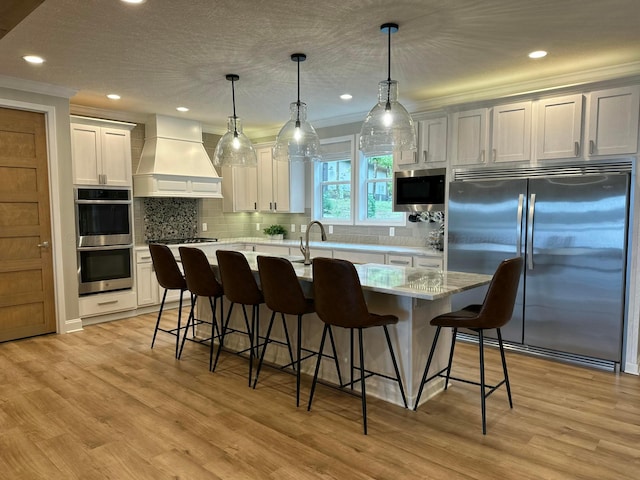 kitchen featuring pendant lighting, custom range hood, appliances with stainless steel finishes, white cabinets, and a kitchen island with sink