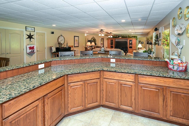 kitchen with dark stone countertops, light tile patterned floors, a ceiling fan, brown cabinetry, and open floor plan