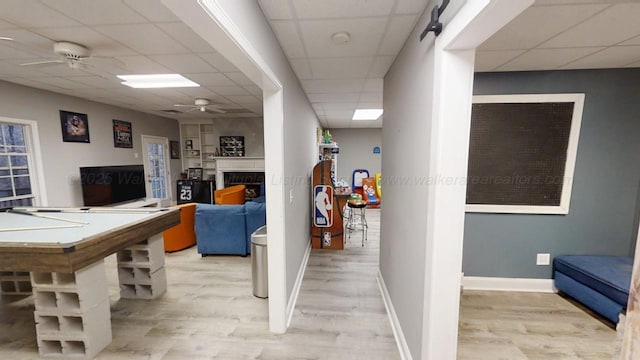 recreation room featuring baseboards, a paneled ceiling, ceiling fan, light wood-type flooring, and a fireplace