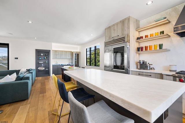 kitchen featuring a kitchen bar, stainless steel built in fridge, wall chimney range hood, light brown cabinetry, and light wood-type flooring