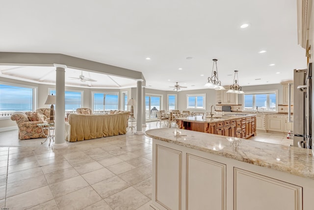 kitchen with hanging light fixtures, ceiling fan, an island with sink, and light stone counters