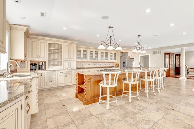 kitchen featuring light stone countertops, decorative light fixtures, decorative columns, an island with sink, and sink