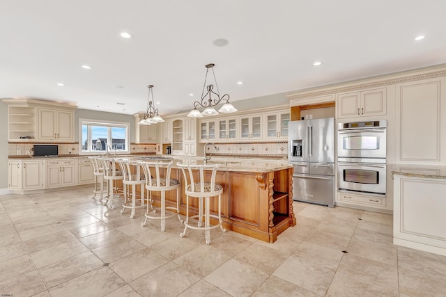 kitchen with appliances with stainless steel finishes, decorative light fixtures, cream cabinetry, an island with sink, and light stone countertops