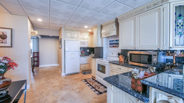 kitchen featuring a paneled ceiling, white appliances, light tile patterned floors, decorative backsplash, and dark stone countertops