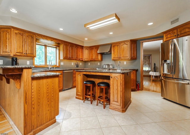 kitchen featuring a kitchen bar, brown cabinetry, appliances with stainless steel finishes, and wall chimney exhaust hood