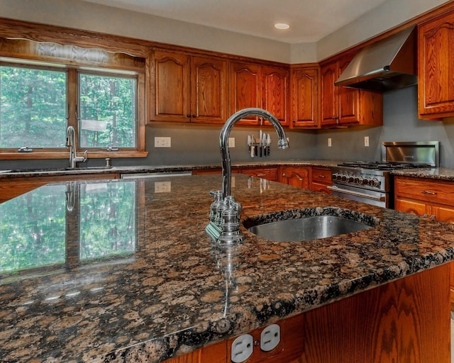 kitchen featuring stainless steel stove, wall chimney range hood, dark stone countertops, and a sink