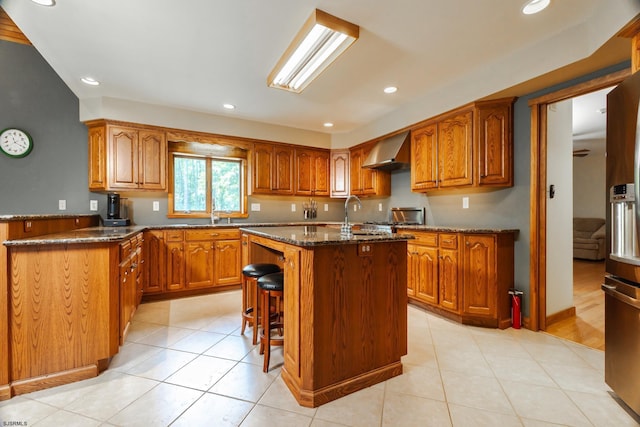 kitchen featuring dark stone countertops, brown cabinetry, a breakfast bar, an island with sink, and wall chimney exhaust hood