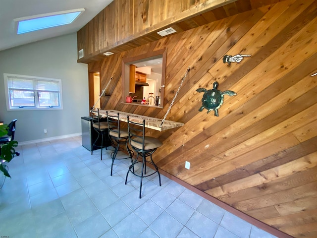 interior space featuring vaulted ceiling with skylight, wooden walls, light tile patterned floors, kitchen peninsula, and a breakfast bar area