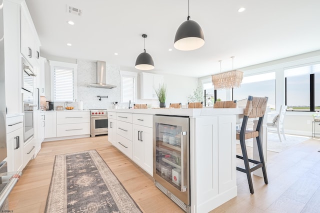 kitchen with light wood-type flooring, stainless steel range oven, white cabinetry, wall chimney range hood, and beverage cooler