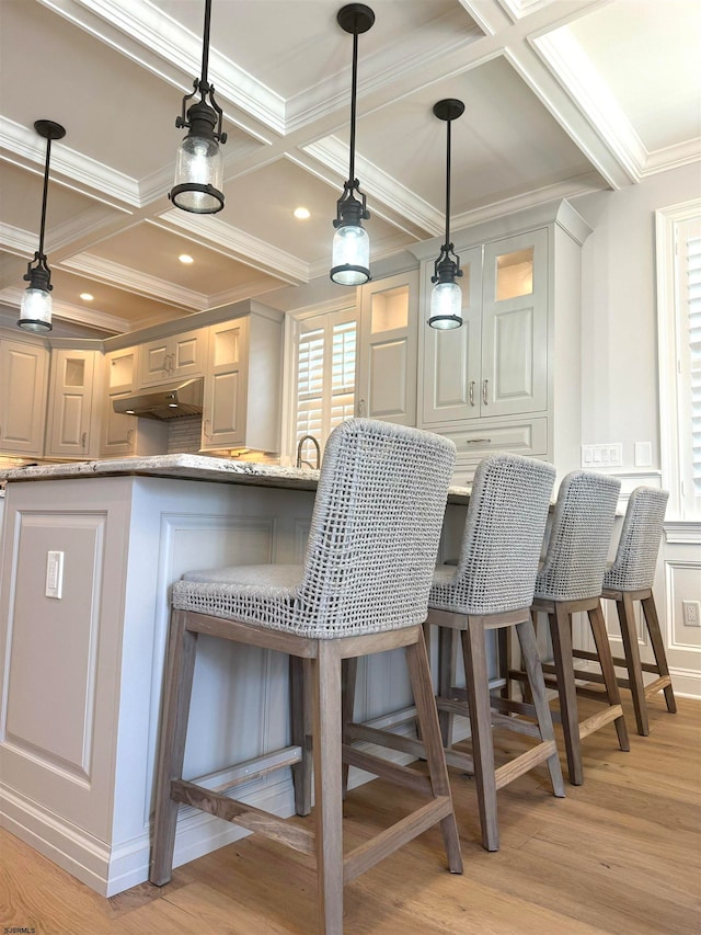 kitchen featuring light wood-type flooring, coffered ceiling, crown molding, light stone countertops, and a breakfast bar