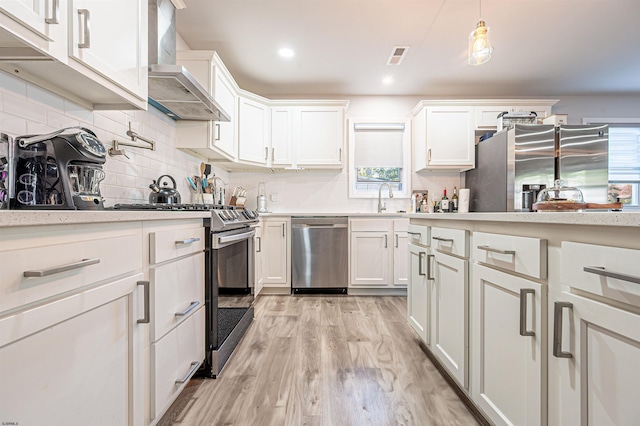 kitchen featuring wall chimney range hood, white cabinetry, light wood-type flooring, decorative light fixtures, and stainless steel appliances