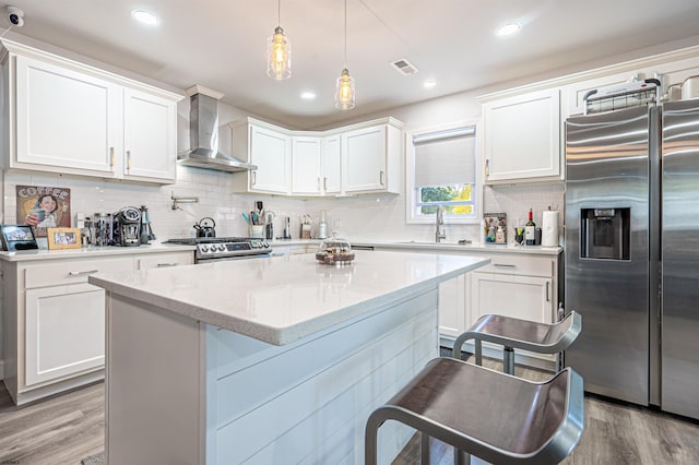 kitchen featuring wall chimney exhaust hood, stainless steel appliances, a kitchen bar, decorative light fixtures, and white cabinetry