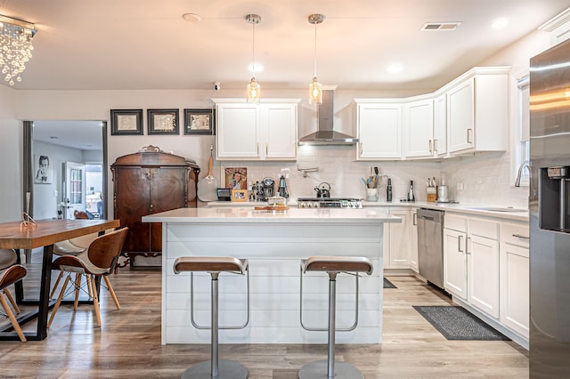 kitchen with wall chimney range hood, appliances with stainless steel finishes, light wood-type flooring, decorative light fixtures, and white cabinets