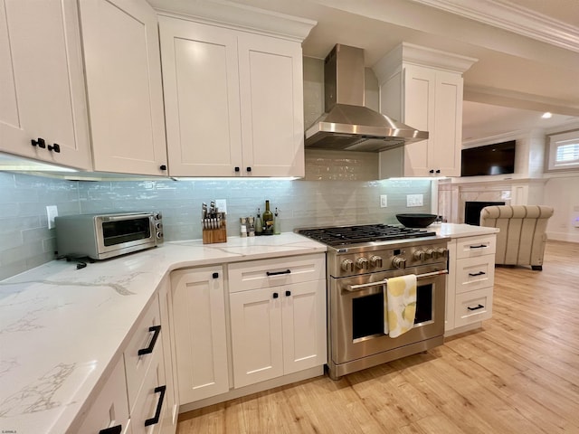 kitchen featuring high end stove, wall chimney range hood, light hardwood / wood-style flooring, ornamental molding, and white cabinets