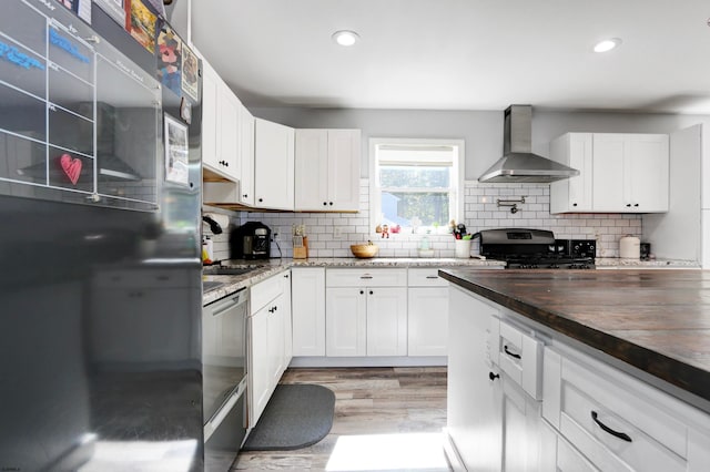 kitchen featuring wall chimney range hood, light hardwood / wood-style flooring, black appliances, white cabinets, and butcher block countertops