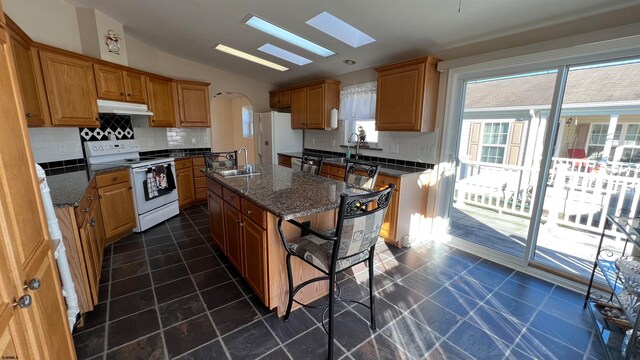 kitchen featuring sink, an island with sink, vaulted ceiling with skylight, backsplash, and white appliances